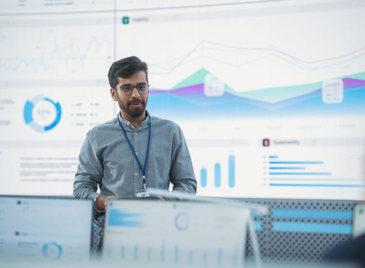 Male Indian Computer Scientist With Tablet Giving Presentation to Diverse Team Of Data Analysts In Front Of Big Digital Screen In Monitoring Room. Colleagues Listening To Training On Automation.